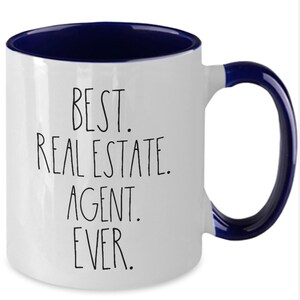 May include: White ceramic coffee mug with a blue rim and handle. The mug has black text that reads "BEST. REAL ESTATE. AGENT. EVER."