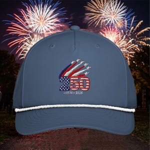 May include: A navy blue baseball cap with a white rope brim. The cap features an embroidered design with the number "250" in red and blue, along with the years "1776-2026". The background shows fireworks.