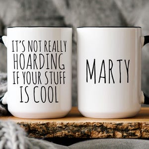 May include: Two white ceramic mugs with black handles and black rims. One mug says "It's not really hoarding if your stuff is cool." The other mug says "Marty".