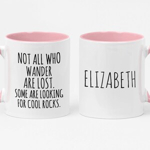 K&ouml;nnte beinhalten: Wei&szlig;e Keramiktasse mit rosafarbenem Rand und Henkel. Die Tasse hat einen schwarzen Text mit der Aufschrift "Not all who wander are lost. Some are looking for cool rocks." Auf der anderen Seite der Tasse steht der Name "Elizabeth" in schwarzer Schrift.