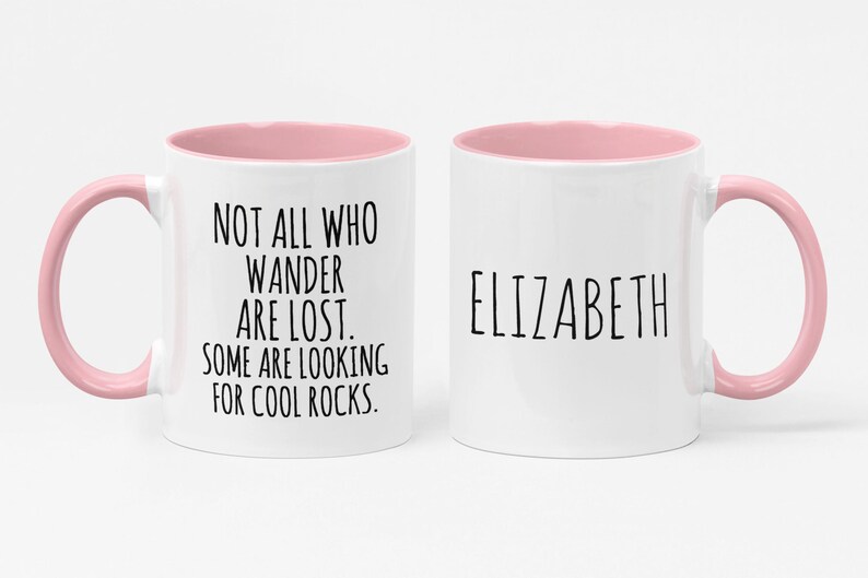 K&ouml;nnte beinhalten: Wei&szlig;e Keramiktasse mit rosafarbenem Rand und Henkel. Die Tasse hat einen schwarzen Text mit der Aufschrift "Not all who wander are lost. Some are looking for cool rocks." Auf der anderen Seite der Tasse steht der Name "Elizabeth" in schwarzer Schrift.