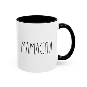 May include: White ceramic mug with a black interior and handle. The word "MAMACITA" is printed in black, handwritten-style font on the front of the mug. The mug is designed for hot or cold beverages.