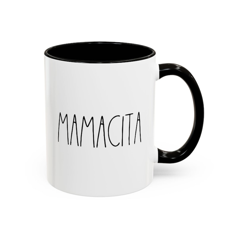 May include: White ceramic mug with a black interior and handle. The word "MAMACITA" is printed in black, handwritten-style font on the front of the mug. The mug is designed for hot or cold beverages.