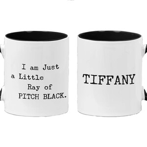 May include: Two white ceramic mugs with black handles and black rims. One mug says "I am Just a Little Ray of PITCH BLACK." The other mug says "TIFFANY".