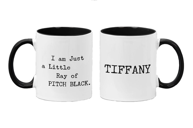 May include: Two white ceramic mugs with black handles and black rims. One mug says "I am Just a Little Ray of PITCH BLACK." The other mug says "TIFFANY".