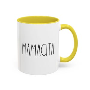 May include: White ceramic mug with a yellow interior and handle. The mug features the word "MAMACITA" in black, handwritten-style lettering. The mug is designed for hot or cold beverages.
