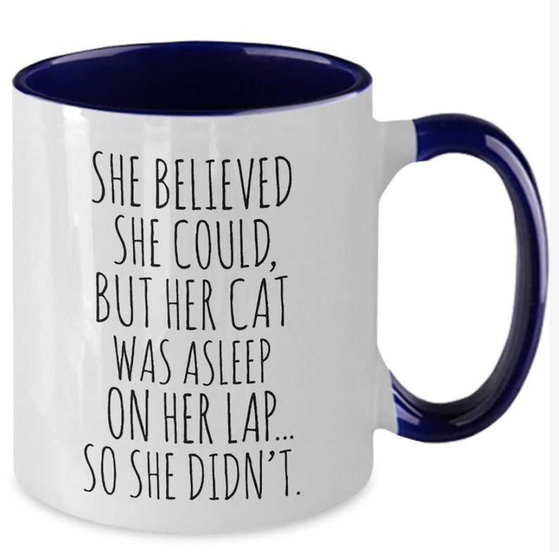 Puede incluir: Taza de caf&eacute; blanca con borde y asa azules. La taza tiene un dicho divertido: "Ella cre&iacute;a que pod&iacute;a, pero su gato estaba dormido en su regazo... as&iacute; que no lo hizo."