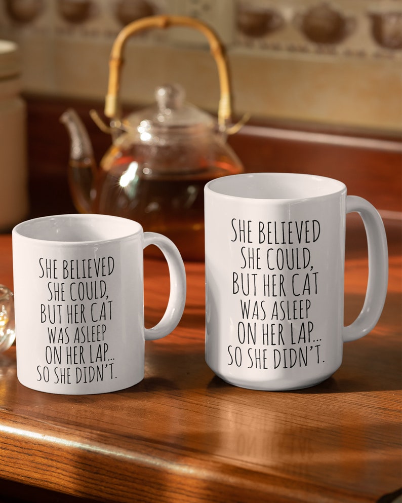 Puede incluir: Dos tazas de cer&aacute;mica blanca con texto negro. El texto en las tazas dice "Ella cre&iacute;a que pod&iacute;a, pero su gato estaba dormido en su regazo... as&iacute; que no lo hizo."