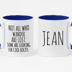 K&ouml;nnte beinhalten: Eine wei&szlig;e Keramik-Kaffeetasse mit blauem Rand und Henkel. Die Tasse hat auf der einen Seite ein Zitat, das lautet "Not all who wander are lost. Some are looking for cool rocks." Auf der anderen Seite der Tasse steht der Name "Jean" in Schwarz.