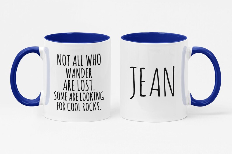 K&ouml;nnte beinhalten: Eine wei&szlig;e Keramik-Kaffeetasse mit blauem Rand und Henkel. Die Tasse hat auf der einen Seite ein Zitat, das lautet "Not all who wander are lost. Some are looking for cool rocks." Auf der anderen Seite der Tasse steht der Name "Jean" in Schwarz.