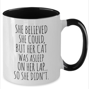 Puede incluir: Taza de caf&eacute; blanca con borde y asa negros. La taza tiene texto negro que dice "SHE BELIEVED SHE COULD, BUT HER CAT WAS ASLEEP ON HER LAP... SO SHE DIDN'T."