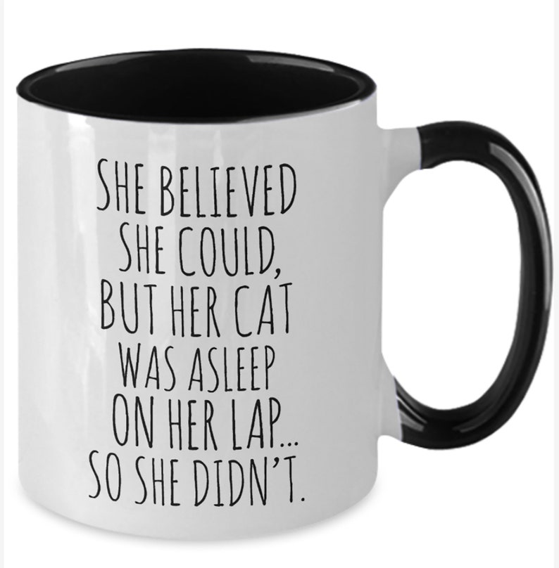 Puede incluir: Taza de caf&eacute; blanca con borde y asa negros. La taza tiene texto negro que dice "SHE BELIEVED SHE COULD, BUT HER CAT WAS ASLEEP ON HER LAP... SO SHE DIDN'T."