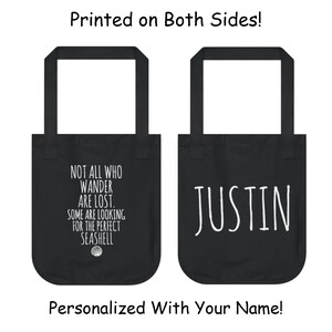 May include: Black tote bag with white text. One side says "Not all who wander are lost. Some are looking for the perfect seashell." The other side says "JUSTIN".