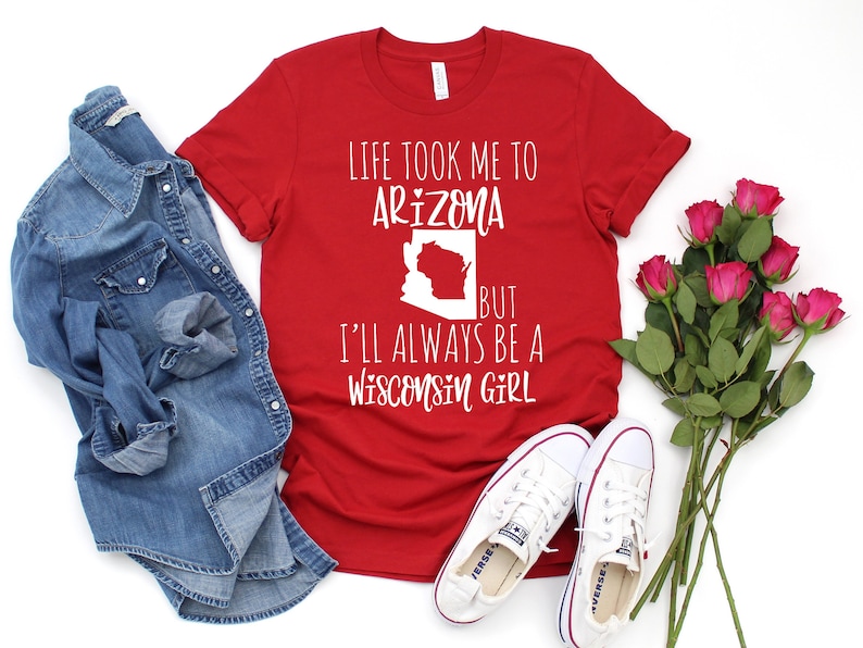 May include: A red t-shirt with white text that reads 'Life took me to Arizona but I'll always be a Wisconsin girl.' The shirt is laid out on a white surface with a pair of white sneakers, a denim jacket, and a bouquet of pink roses.