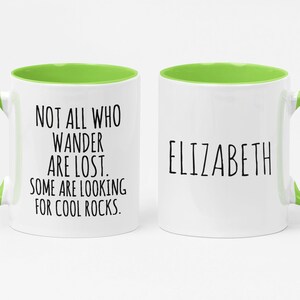 K&ouml;nnte beinhalten: Wei&szlig;e Keramiktasse mit gr&uuml;nem Rand und Henkel. Die Tasse hat schwarzen Text mit der Aufschrift "Not all who wander are lost. Some are looking for cool rocks." Die andere Seite der Tasse tr&auml;gt den Namen "Elizabeth" in schwarzer Schrift.