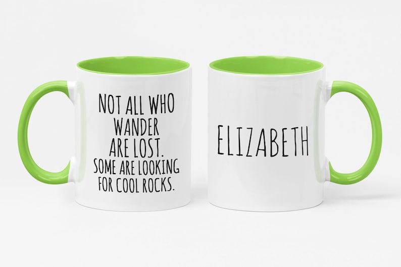 K&ouml;nnte beinhalten: Wei&szlig;e Keramiktasse mit gr&uuml;nem Rand und Henkel. Die Tasse hat schwarzen Text mit der Aufschrift "Not all who wander are lost. Some are looking for cool rocks." Die andere Seite der Tasse tr&auml;gt den Namen "Elizabeth" in schwarzer Schrift.