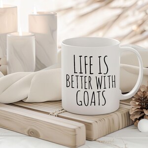 May include: White ceramic coffee mug with black text that reads "Life is better with goats". The mug is sitting on a wooden tray with a white cloth and pine cones.