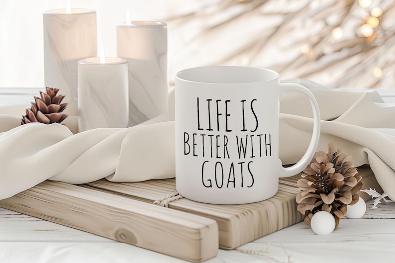 May include: White ceramic coffee mug with black text that reads "Life is better with goats". The mug is sitting on a wooden tray with a white cloth and pine cones.