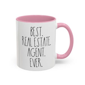 May include: White ceramic coffee mug with a pink rim and handle. The mug has black text that reads "BEST. REAL ESTATE. AGENT. EVER."