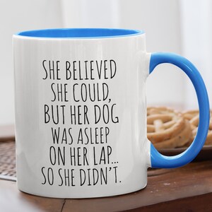 Op de afbeelding: Een witte keramische koffiemok met een blauwe rand en handvat. De mok heeft zwarte tekst met de tekst "Ze geloofde dat ze kon, maar haar hond sliep op haar schoot... dus deed ze het niet."
