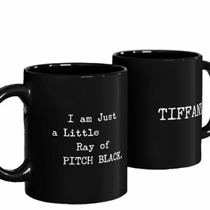 May include: Two black ceramic mugs with white text. One mug says "I am just a little Ray of PITCH BLACK." The other mug says "TIFFANY".