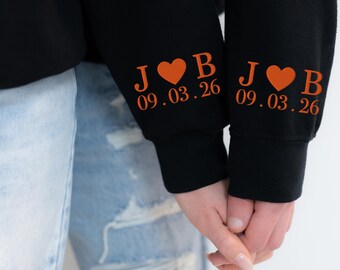 Könnte beinhalten: Zwei schwarze Langarm-Sweatshirts mit orangefarbenem Text und Grafiken an den Ärmelbündchen. Der Aufdruck zeigt die Buchstaben J und B, getrennt durch ein Herzsymbol, mit dem Datum 09.03.26 direkt darunter in einer fetten Serifenschrift.