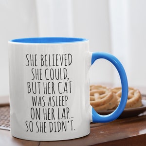 Puede incluir: Taza de caf&eacute; de cer&aacute;mica blanca con borde y asa azules. La taza tiene un dicho divertido impreso: "Ella cre&iacute;a que pod&iacute;a, pero su gato estaba dormido en su regazo... as&iacute; que no lo hizo."