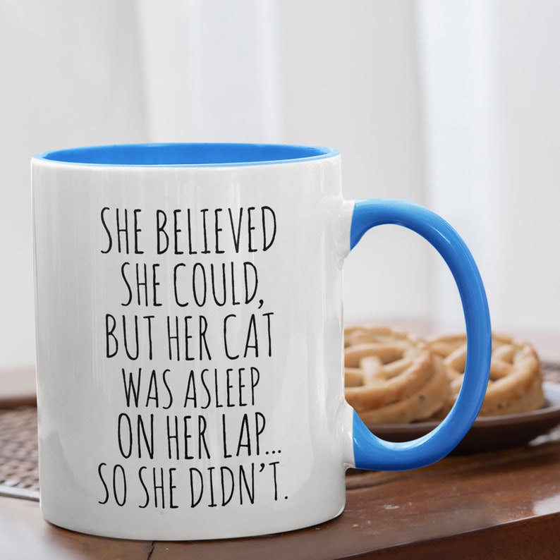 Puede incluir: Taza de caf&eacute; de cer&aacute;mica blanca con borde y asa azules. La taza tiene un dicho divertido impreso: "Ella cre&iacute;a que pod&iacute;a, pero su gato estaba dormido en su regazo... as&iacute; que no lo hizo."