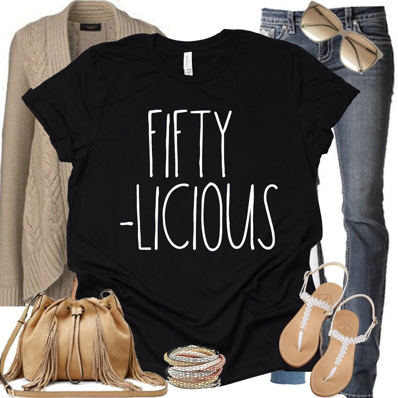 May include: Black t-shirt with the text "FIFTY -LICIOUS" in white. The shirt is paired with a beige cardigan, blue jeans, a brown purse, and bracelets.