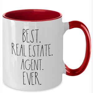 May include: White ceramic coffee mug with a red rim and handle. The mug has black text that reads "Best. Real Estate. Agent. Ever."