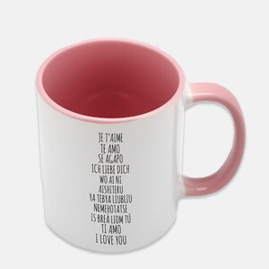 May include: White ceramic mug with a pink rim and handle. The mug has black text that reads "Je t'aime, Te amo, Se agapo, Ich liebe dich, Wo ai ni, Aishiteru, Ya tebya liubliu, Nemehotatse, Is brea liom tu, Ti amo, I love you."