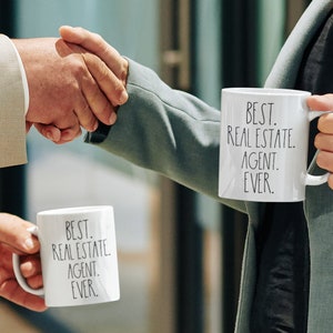 May include: Two people shaking hands while holding white mugs with the text "BEST. REAL ESTATE. AGENT. EVER." on them.