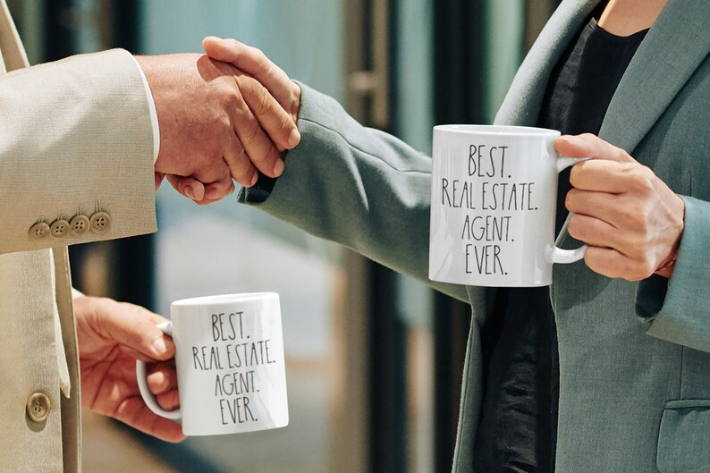 May include: Two people shaking hands while holding white mugs with the text "BEST. REAL ESTATE. AGENT. EVER." on them.