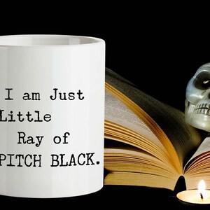 May include: White ceramic mug with black text that reads "I am Just a Little Ray of PITCH BLACK." The mug is next to a book and a skull with a lit candle.