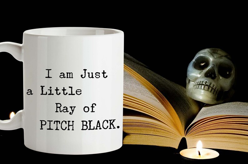 May include: White ceramic mug with black text that reads "I am Just a Little Ray of PITCH BLACK." The mug is next to a book and a skull with a lit candle.