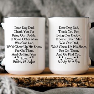 May include: Two white ceramic mugs with black handles and black rims. The mugs have a black paw print design and text that reads "Dear Dog Dad, Thank You For Being Our Daddy. If Some Other Man Was Our Dad, We'd Chew Up His Shoes, Pee On Them, And Go Find You. Love, Buddy & Aqua"