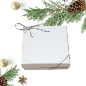May include: A white gift box with a silver ribbon tied in a bow. The box is surrounded by pine branches and gold ornaments.