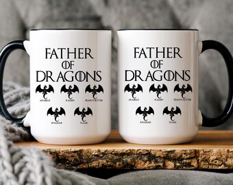 May include: Two white ceramic mugs with black handles and rims. The text "FATHER OF DRAGONS" is printed in black, with a circular icon replacing the "O". Below, five black dragon silhouettes are labeled with the names Arabelle, Kaiden, Peanut Butter, Rhaegar, and Flame.