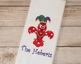 May include: White kitchen towel with an embroidered red crawfish wearing a purple and green jester hat and a purple and gold beaded necklace. The text "The Heberts" is embroidered below the crawfish.