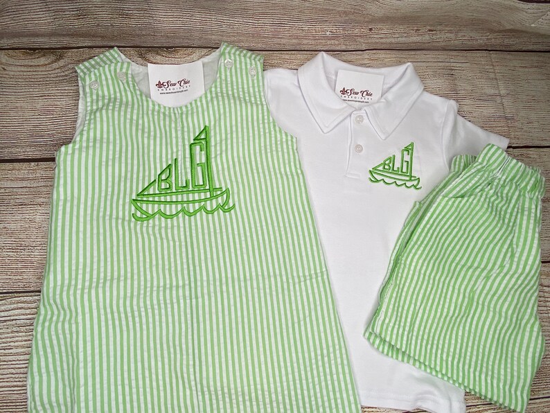 May include: A green and white striped outfit for a baby or toddler. The outfit includes a sleeveless romper, a short-sleeved polo shirt, and shorts. The romper and shorts have a sailboat design with the initials "BLG" embroidered on them. The polo shirt has the same sailboat design embroidered on the front.