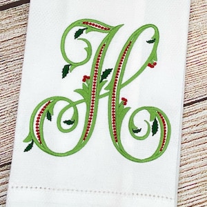 May include: A white embroidered kitchen towel with a green and red holly design featuring the letter "H" in a cursive font.