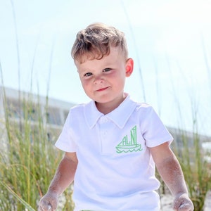 May include: A young boy wearing a white polo shirt with a green sailboat graphic on the front. The shirt has the letters "ALNI" on the sailboat. The boy is standing on a sandy beach with tall green grass in the background.