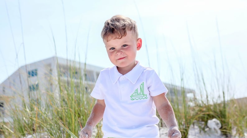 May include: A young boy wearing a white polo shirt with a green sailboat graphic on the front. The shirt has the letters "ALNI" on the sailboat. The boy is standing on a sandy beach with tall green grass in the background.