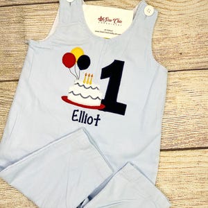 May include: Light blue baby romper with a birthday cake and balloons applique. The number 1 is appliqued in black, and the name "Elliot" is embroidered below. The romper has button closures on the shoulders.