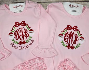 May include: Two light pink long-sleeve shirts with ruffled cuffs and hems. Each shirt features an embroidered Christmas wreath design with red bows, green leaves, and two Santa Claus faces. The left shirt includes the text First Christmas.