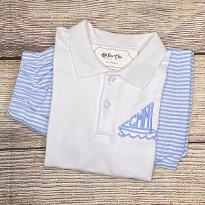 May include: White polo shirt with a blue sailboat embroidered on the front. The sailboat has the letters "GMM" on the sail. The shirt is folded and laid on a wooden surface.