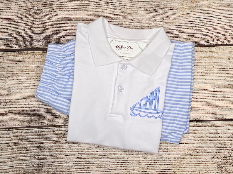 May include: White polo shirt with a blue sailboat embroidered on the front. The sailboat has the letters "GMM" on the sail. The shirt is folded and laid on a wooden surface.