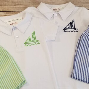 May include: Two white polo shirts with short sleeves and green and black embroidered sailboats with initials. The shirts are paired with striped shorts, one green and one blue.