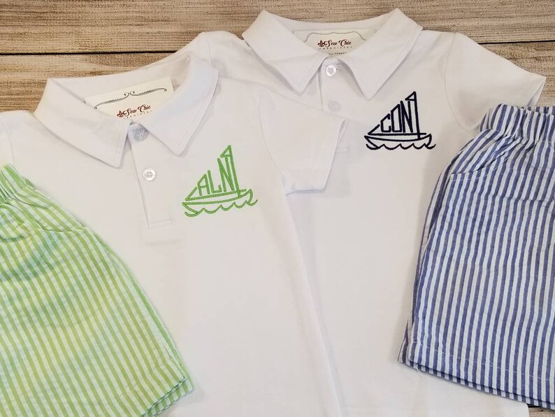 May include: Two white polo shirts with short sleeves and green and black embroidered sailboats with initials. The shirts are paired with striped shorts, one green and one blue.