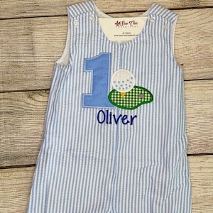May include: Blue and white striped seersucker romper with a golf-themed applique. The applique features the number 1, a golf ball, and a green golf course design. The name "Oliver" is embroidered below the design.